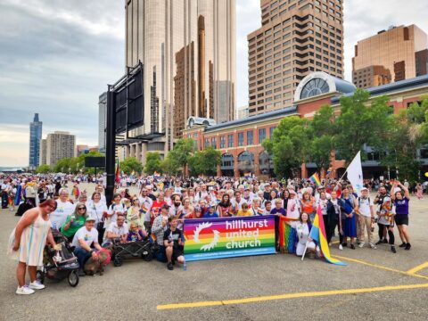 A church congregation gathers in a crowd. They are holding up a rainbow sign that says Hillhurst United Church.