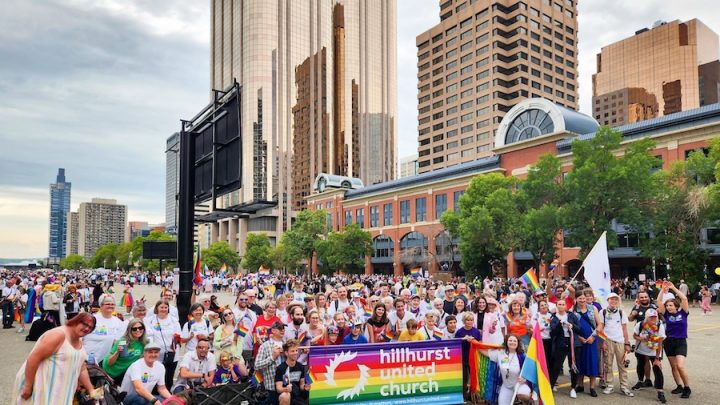 A church congregation gathers in a crowd. They are holding up a rainbow sign that says Hillhurst United Church.