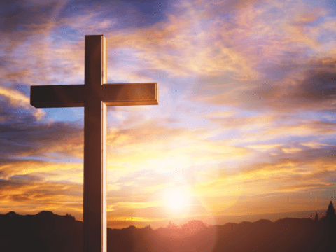 Photograph of a cross against a sunset. The sky is blue with pink, purple and white clouds. The sun is shining