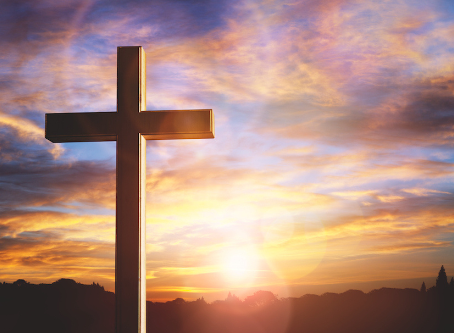 Photograph of a cross against a sunset. The sky is blue with pink, purple and white clouds. The sun is shining