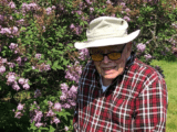 An elderly white man in a beige bucket hat and red and white flannel shirt poses in front of a flowering bush. He is wearing black-framed glasses and smiling.