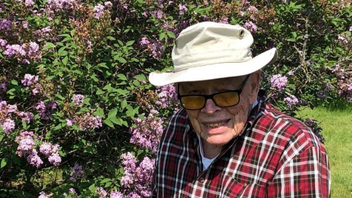 An elderly white man in a beige bucket hat and red and white flannel shirt poses in front of a flowering bush. He is wearing black-framed glasses and smiling.