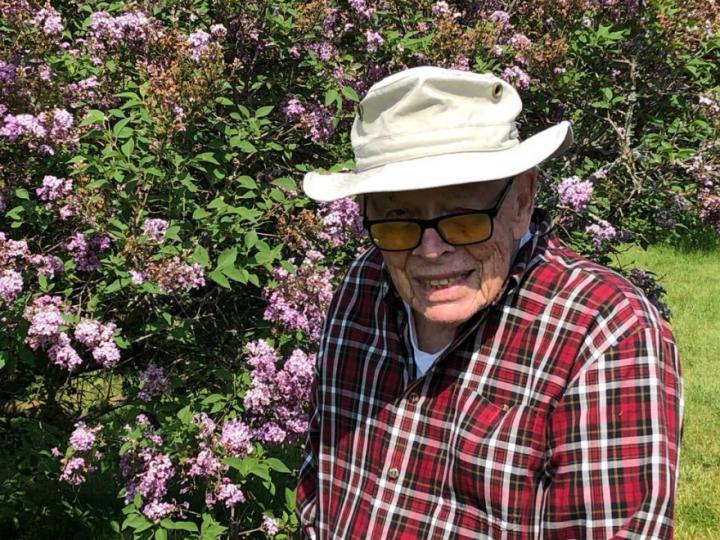 An elderly white man in a beige bucket hat and red and white flannel shirt poses in front of a flowering bush. He is wearing black-framed glasses and smiling.