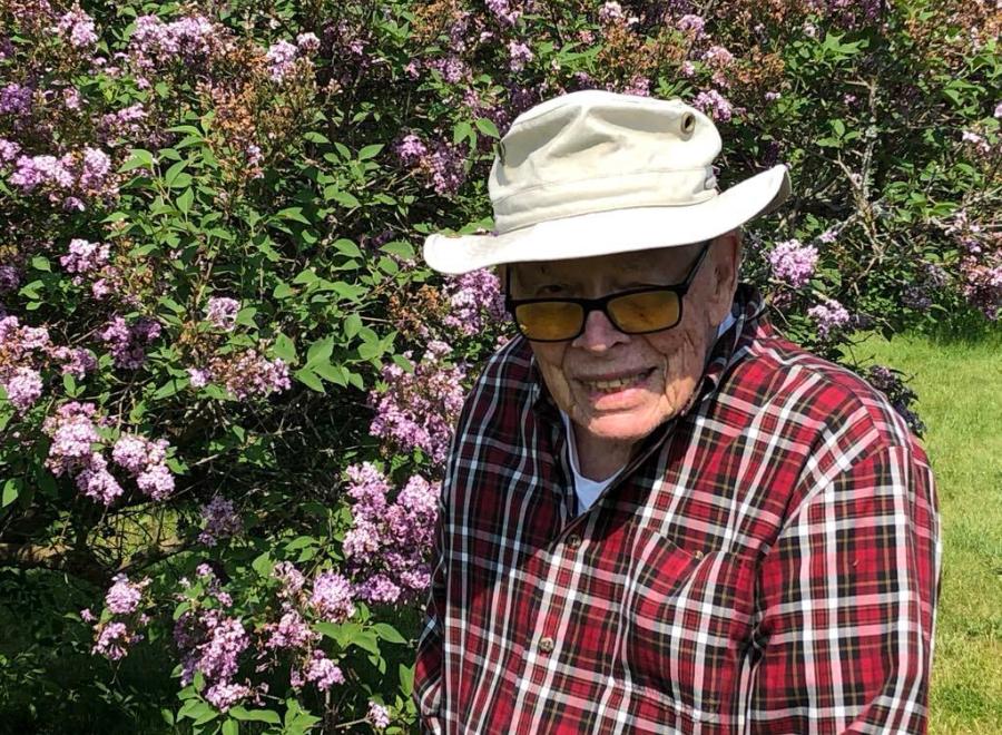An elderly white man in a beige bucket hat and red and white flannel shirt poses in front of a flowering bush. He is wearing black-framed glasses and smiling.