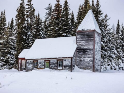 A wooden church stands in front of a snowy landscape. There are pine trees in the background, also covered in snow