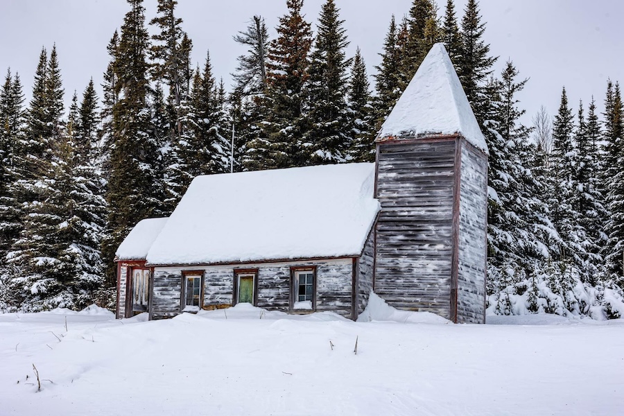 A wooden church stands in front of a snowy landscape. There are pine trees in the background, also covered in snow