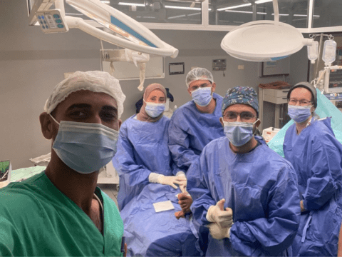 A medical team of five people in blue face masks, scrubs and caps pose in a hospital operating room
