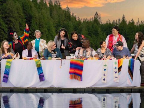 People of a variety of ethnicities and ages sit at and stand around a table covered with a white cloth on a dock