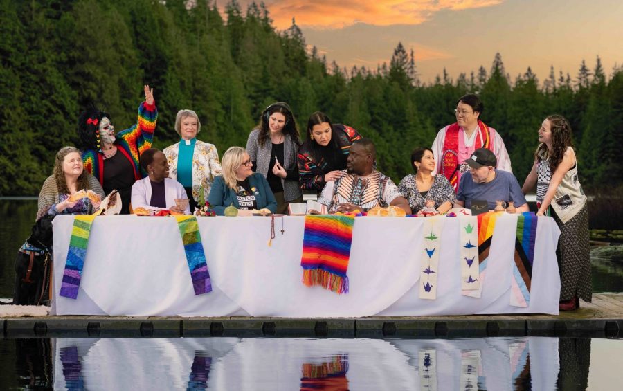 People of a variety of ethnicities and ages sit at and stand around a table covered with a white cloth on a dock
