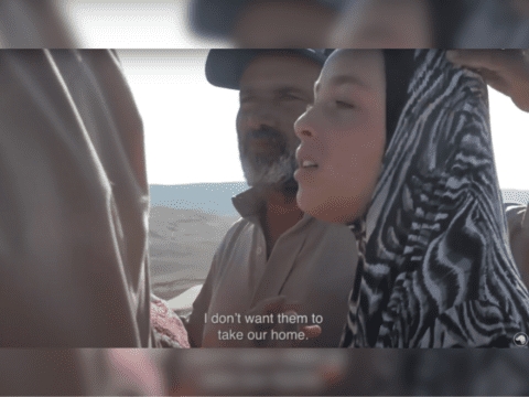 A brown-skinned man with a white beard and cap stands next to a hijabi woman. She is saying, "I don't want them to take my home," in subtitles.