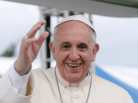 An older white man in a papal outfit with white cap, white button-up robe and a necklace smiles and has his hand up waving
