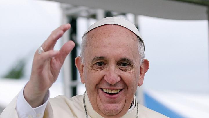 An older white man in a papal outfit with white cap, white button-up robe and a necklace smiles and has his hand up waving