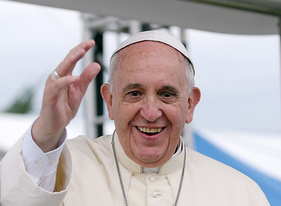 An older white man in a papal outfit with white cap, white button-up robe and a necklace smiles and has his hand up waving