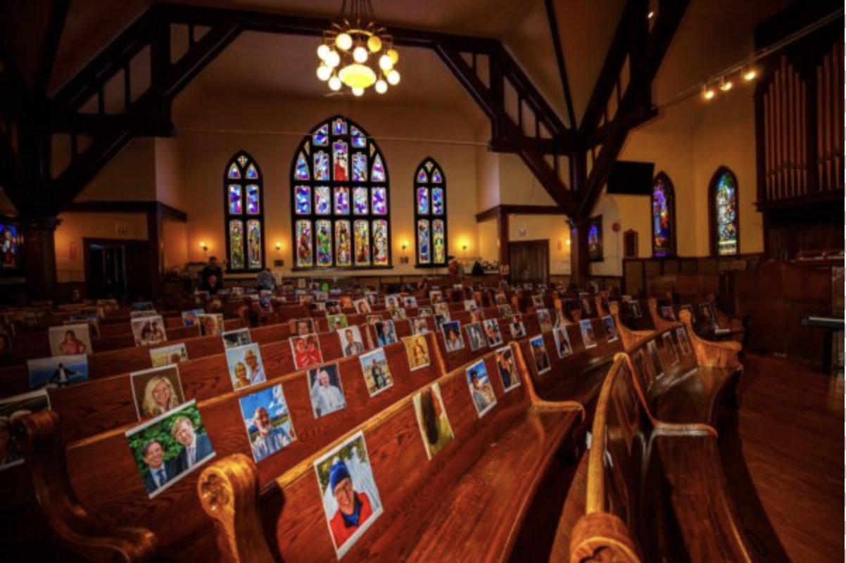Empty pews in a church have photos of congregants on them. There are stained glass windows and beams from the church's structure.