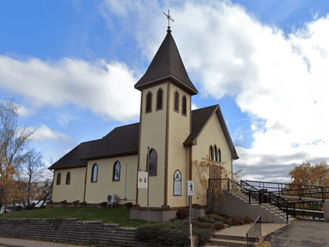 A yellow church with a brown roof stands before blue skies and white clouds