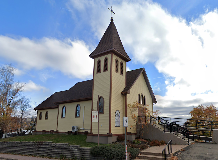 A yellow church with a brown roof stands before blue skies and white clouds