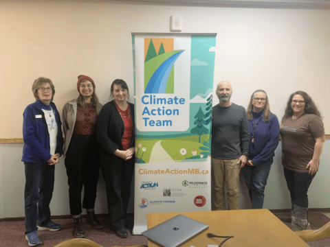 A group of white people, both men and women, stand around a sign that says "Climate Action Team" in blue. There is a silver laptop on a table in front of the sign. The people are of different ages.