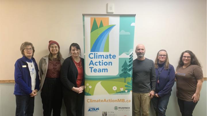 A group of white people, both men and women, stand around a sign that says "Climate Action Team" in blue. There is a silver laptop on a table in front of the sign. The people are of different ages.