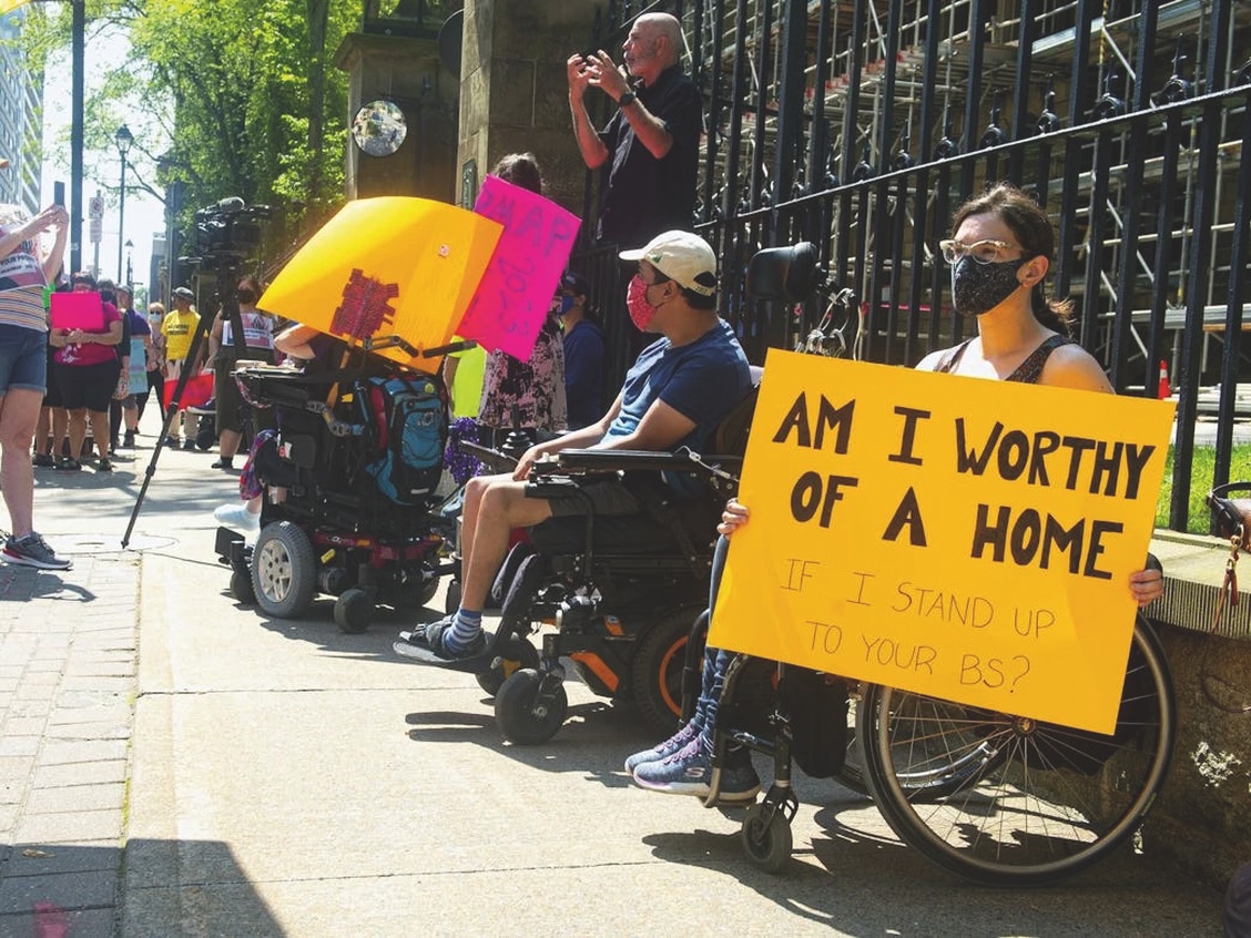 A number of disability justice activists in wheelchairs and masks are in front of an iron gate. One of them holds an orange sign that says" Am I worth of a home if I stand up to your BS"