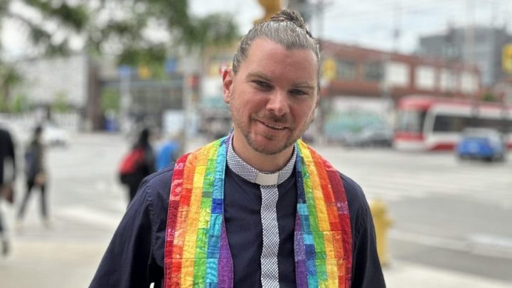 A young white man poses on the street. His hair is pulled back in a bun and he is wearing a black and rainbow-coloured minister's shirt with a collar
