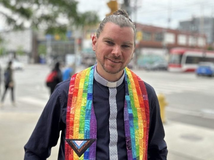 A young white man poses on the street. His hair is pulled back in a bun and he is wearing a black and rainbow-coloured minister's shirt with a collar