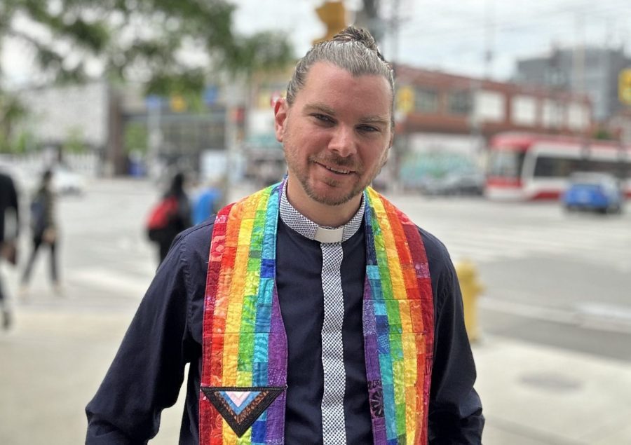 A young white man poses on the street. His hair is pulled back in a bun and he is wearing a black and rainbow-coloured minister's shirt with a collar