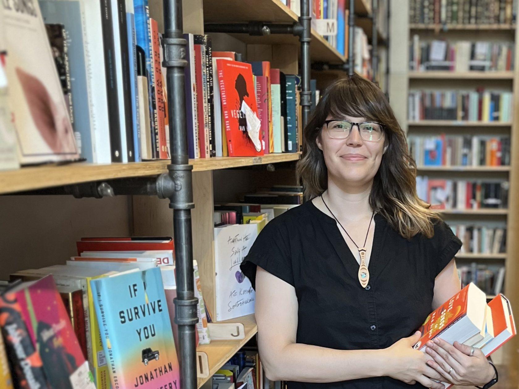 An Indigenous woman with brown hair, brown skin and black-framed glasses stands in front of a bookshelf