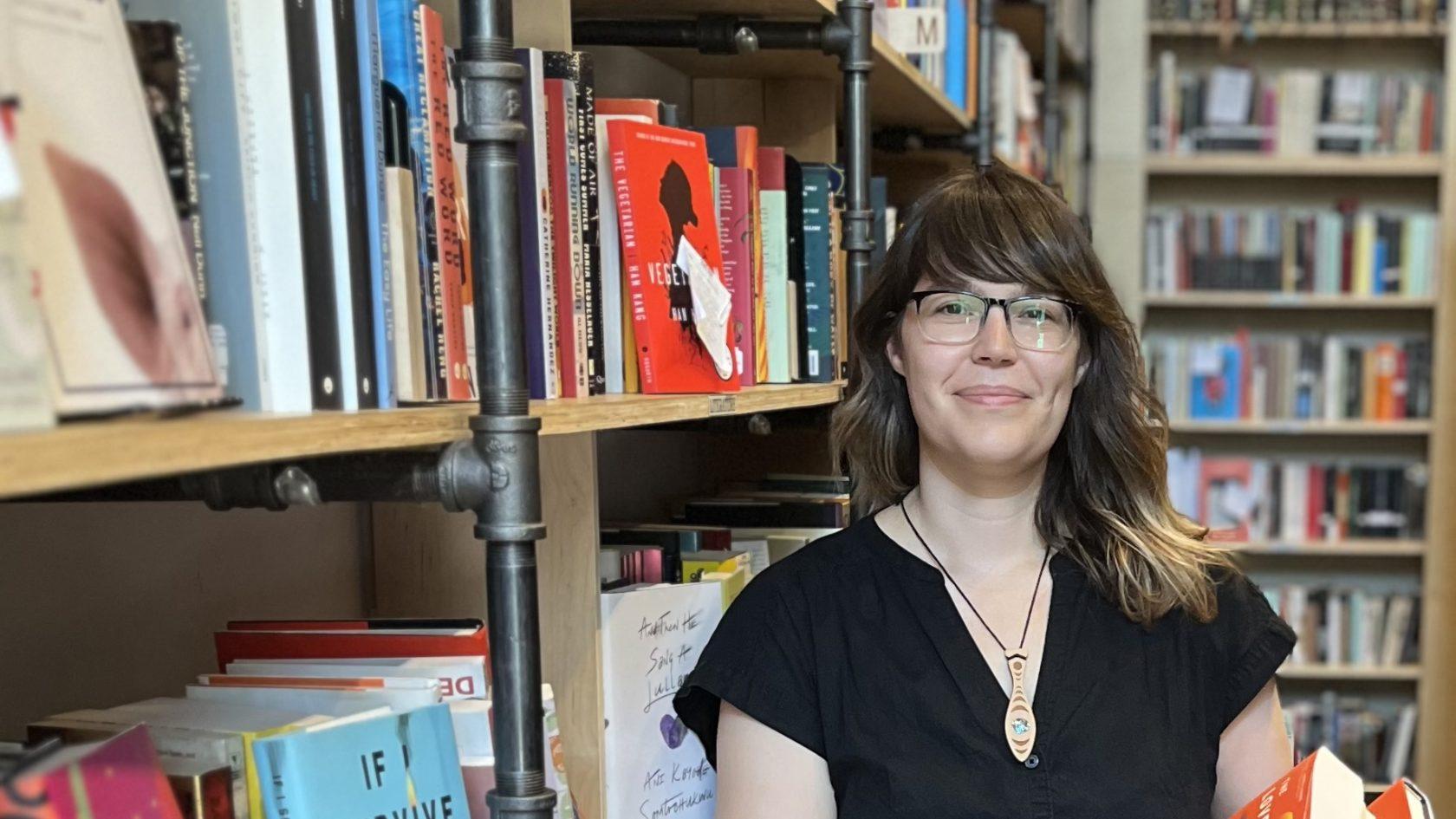 An Indigenous woman with brown hair, brown skin and black-framed glasses stands in front of a bookshelf