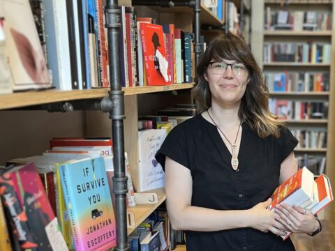 An Indigenous woman with brown hair, brown skin and black-framed glasses stands in front of a bookshelf
