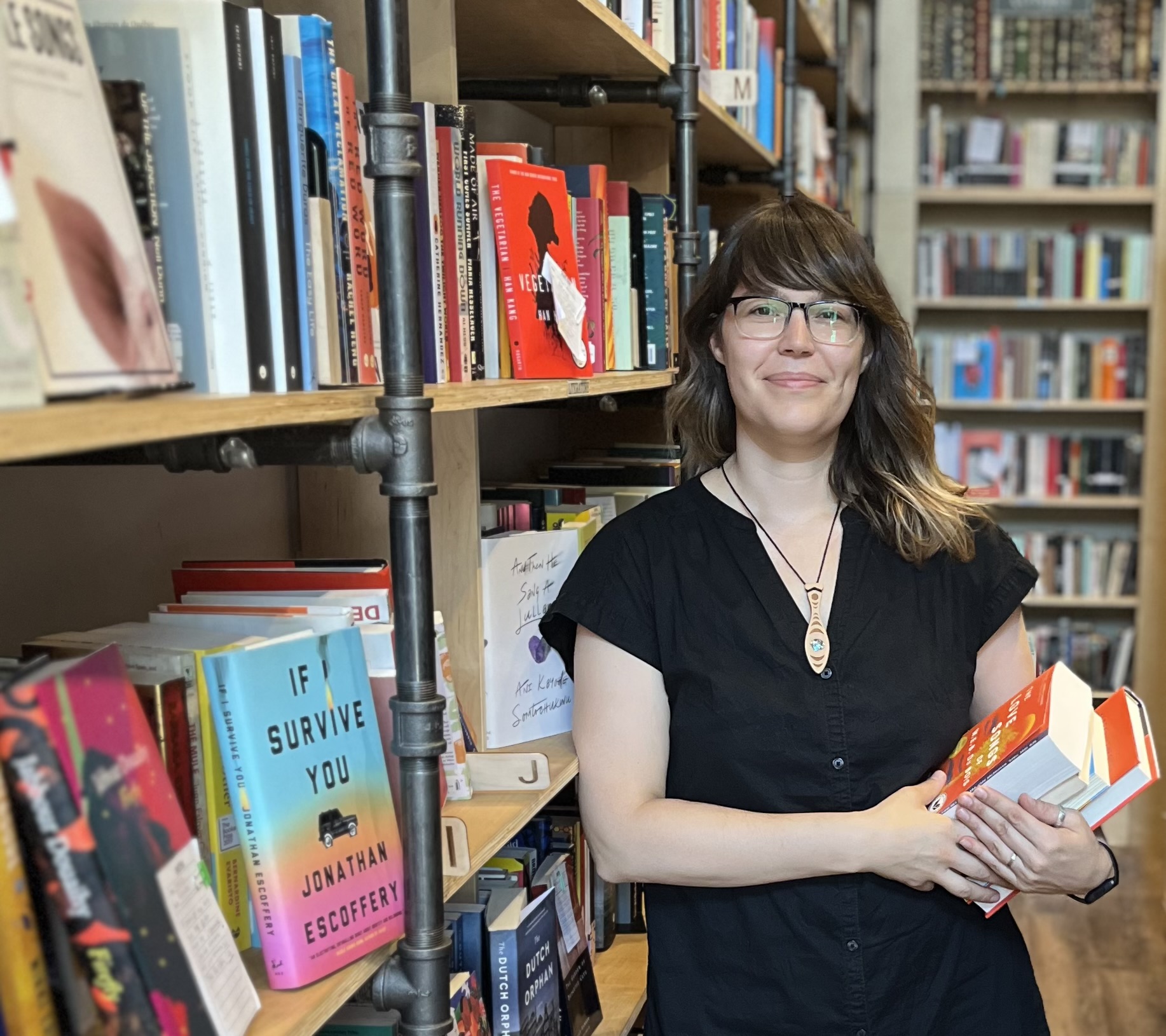 An Indigenous woman with brown hair, brown skin and black-framed glasses stands in front of a bookshelf