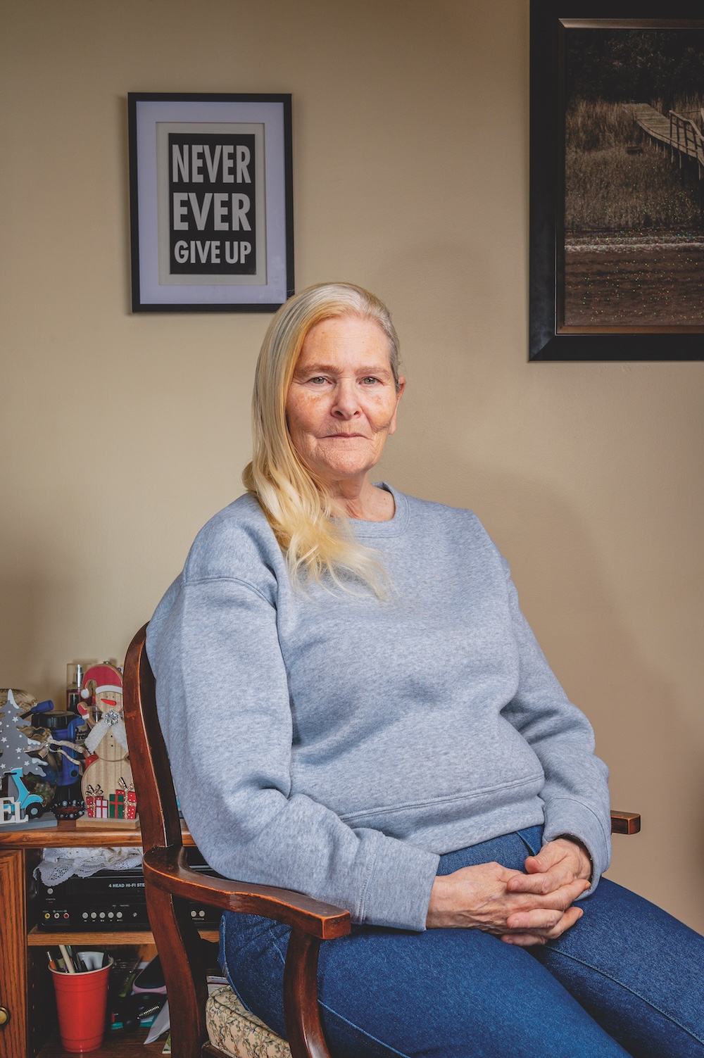 An older white woman with white hair sits in a chair looking at the camera. She is wearing a blue sweatshirt and jeans.