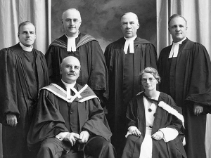 Black and white photo of five men and one woman posing. The four men in the back are standing and one man and woman are in the front row sitting. They are all wearing robes. 