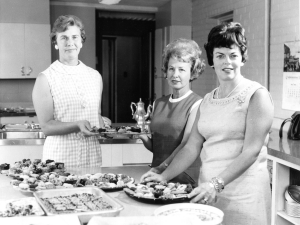 A black and white photo of three women in sleeveless dresses. One of them is holding a tray of baked goods. She has short hair, earrings and glasses on. A shorter woman stands next to her and helps hold the tray. A woman on the right has brown hair and is wearing pearls, a broach, a ring and a watch and bracelet. On the table in front of them are several trays of baked goods and plates. 