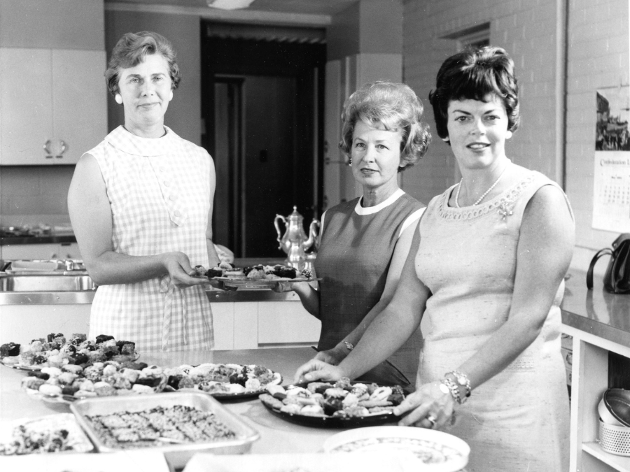 A black and white photo of three women in sleeveless dresses. One of them is holding a tray of baked goods. She has short hair, earrings and glasses on. A shorter woman stands next to her and helps hold the tray. A woman on the right has brown hair and is wearing pearls, a broach, a ring and a watch and bracelet. On the table in front of them are several trays of baked goods and plates.