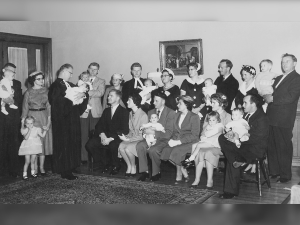 Black and white photo of several people posing. The men are dressed in suits and the women are in dresses. There is a front row of six people sitting. The adults are holding babies and one man on the left is in a black robe holding a child. 