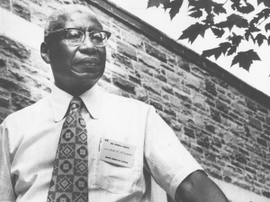 A black and white photo of a Black man with white hair in glasses, short-sleeve button up shirt and a tie. He is wearing a name tag that says Wibur Howard. He is posing in front of a brick wall.