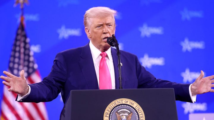 Donald Trump speaking at a podium with arms outstretched. Blue CPAC backdrop and American flag behind him.