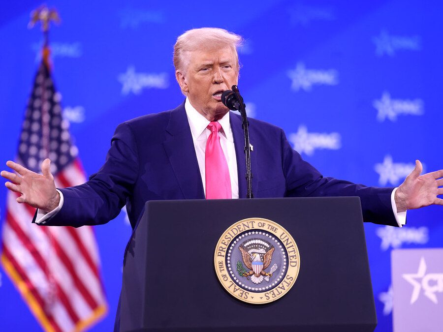 Donald Trump speaking at a podium with arms outstretched. Blue CPAC backdrop and American flag behind him.