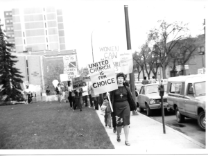 Black and white photo of women marching. In the front is a woman in a black skirt, shirt and cardigan. She is wearing a cross and holding a sign that says, "United Church is for choice"