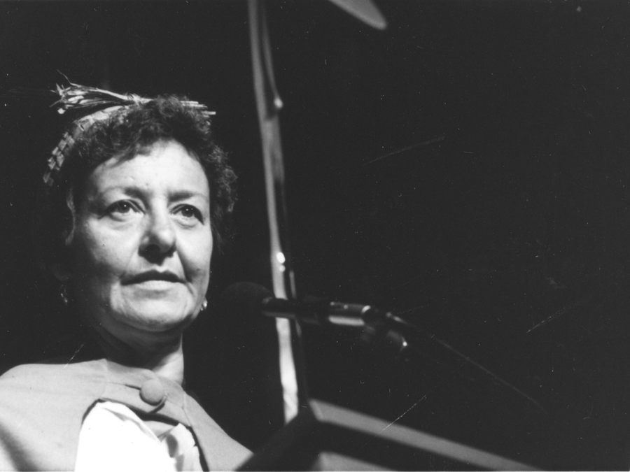 A black and white image of an Indigenous woman with curly hair and a ribbon in her hair. She stands before a microphone