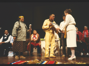 An Indigenous man with braids holds a staff. He is standing next to another Indigenous man who is presenting a framed copy of an apology to a white woman with glasses, short hair and a white shirt, skirt and heels. Behind them are several Indigenous people seated and looking on