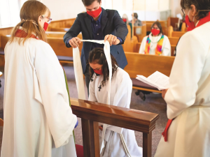 A man in a black suit and red face mask puts a white ribbon over a girl who is kneeling. She is wearing a black cross and white robes. In front of her stands two women in white robes and red face masks. The woman on the right is holding a book. 
