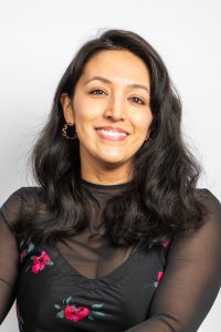 A brown-skinned woman with dark hair smiles. She is wearing a black shirt with red flowers on it and gold-hooped earrings