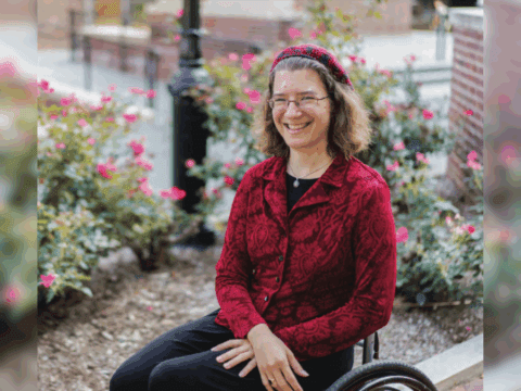 A white woman with curly hair is in a wheelchair and smiling. She has glasses and is wearing a red hat and red button-up shirt, black pants. Her hands are in her lap.