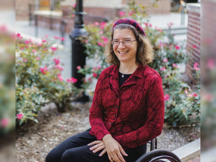 A white woman with curly hair is in a wheelchair and smiling. She has glasses and is wearing a red hat and red button-up shirt, black pants. Her hands are in her lap.