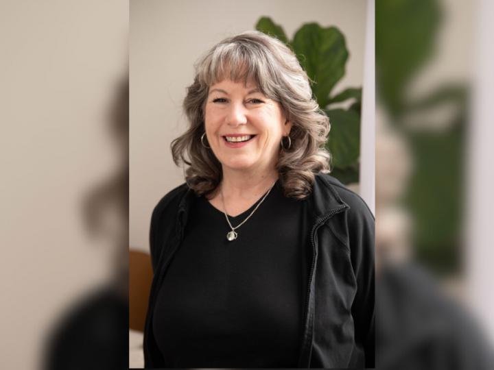 A middle-aged white woman with brown and grey hair poses smiling. She is wearing a black shirt and cardigan with a necklace and earrings on