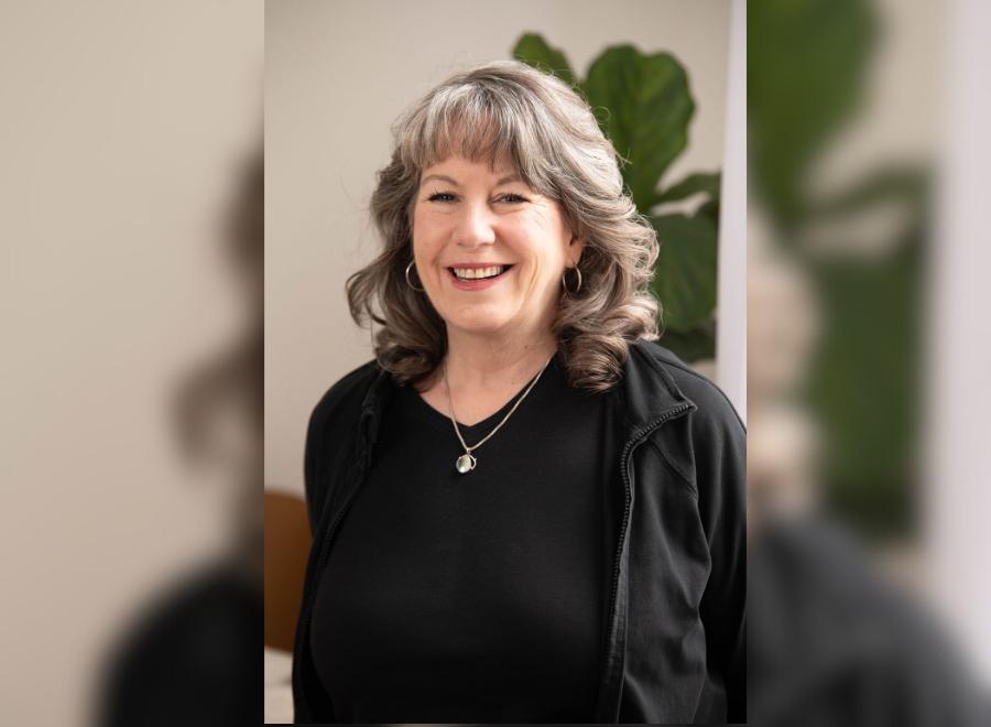 A middle-aged white woman with brown and grey hair poses smiling. She is wearing a black shirt and cardigan with a necklace and earrings on