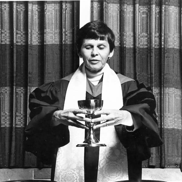 Black and white image of a woman wearing robes and holding a chalice. She has short hair and is wearing earrings