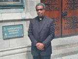 A middle-aged Black man with brown skin, salt-and-pepper curly hair stands in front of church doors. He is dressed in a black pastor's outfit and black shoes. He is holding his hands together in front of him.