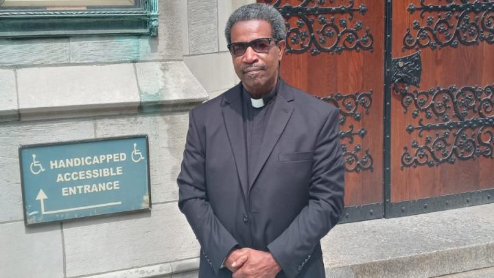 A middle-aged Black man with brown skin, salt-and-pepper curly hair stands in front of church doors. He is dressed in a black pastor's outfit and black shoes. He is holding his hands together in front of him.